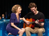 Malu Valle e Gil Coelho rindo no sofá azul do cenário de Matilde, ele tocando violão